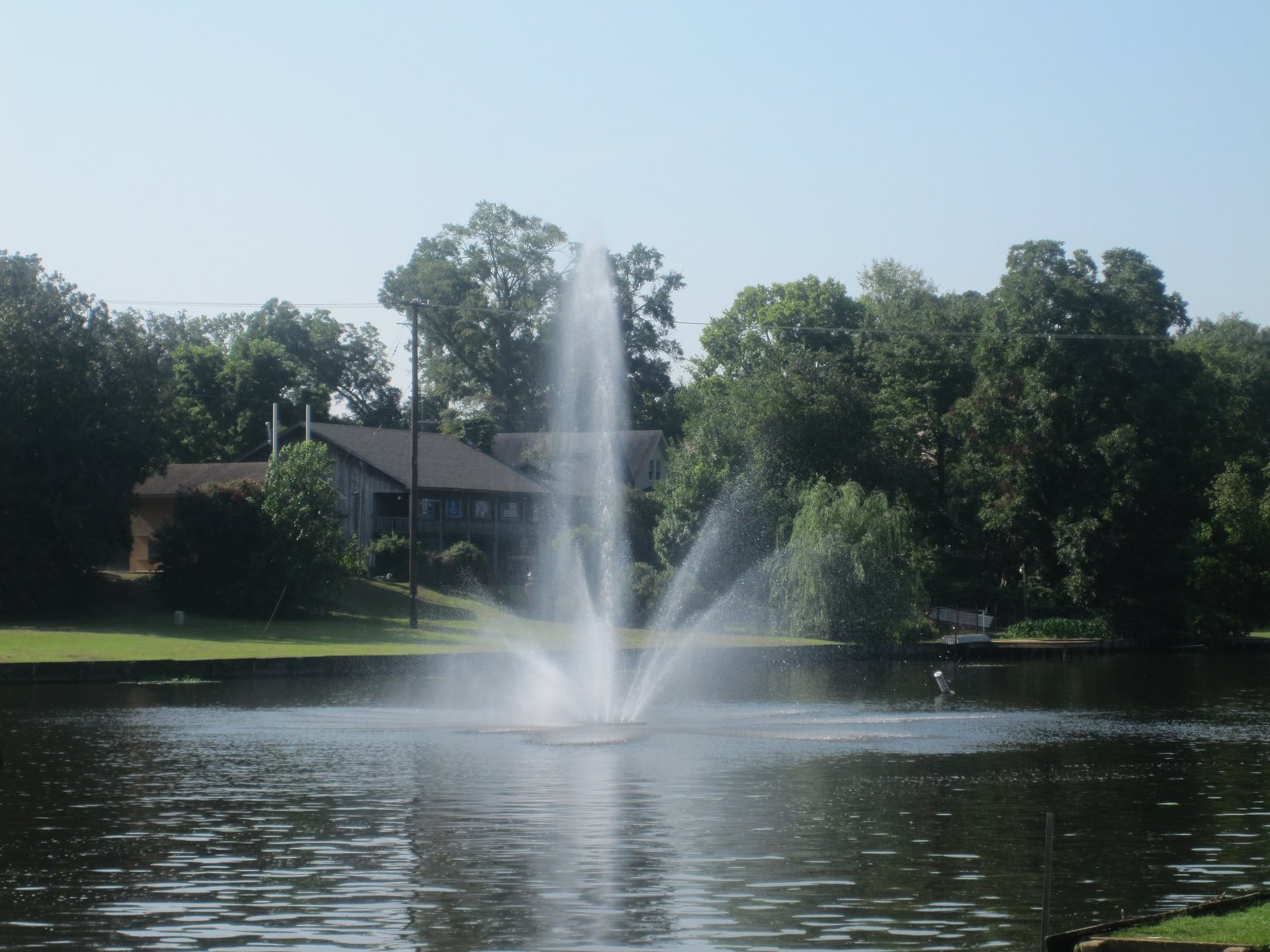 A fountain spraying in Cane River Lake in downtown Natchitoches, with a house among the trees on the far bank